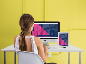 Woman looking at an iMac computer and tablet at a white desk with a yellow background